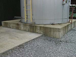 This image shows a grey metal storage tank sitting on a cracked concrete foundation. The foundation has a very large transverse crack running along the width of the foundation, parallel to the ground. Areas of the foundation are stained.