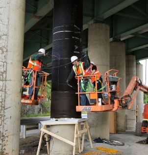 This photo shows three men on a mechanical lift applying a wire mesh around a cylindrical concrete column. The column is already wrapped with a thick, black fabric.