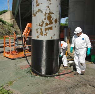 This image shows a two men standing next to a grey concrete column. One man is wrapping the base of the column with a black mesh fabric.