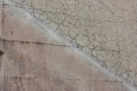 This image shows a close up of the surface of a concrete bridge wall. A 'line' can be seen running diagonally across the image, where the bridge was recently excavated. The top part of the diagonal, which is the section where the structure was exposed to the environment, shows extensive map cracking. Below the diagonal, which is the section where the structure was buried, shows no sign of cracking.