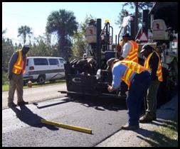 A paving crew is pictured checking the pavement with a level and the crew chief gives a 'thumbs up' to signal that the newly paved section is level.