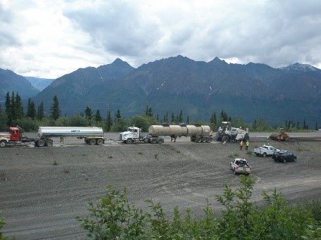 In-place recycling train performing full-depth reclamation. Lead truck is a water truck, followed by an emulsion truck. A reclaimer is processing the existing pavement and followed by a roller.