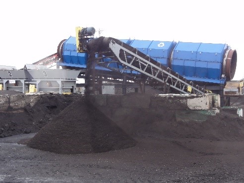 This is a trommel, a rotating cylinder with small openings, that is sorting the properly sized recycled asphalt shingles onto a conveyor which is making a small stockpile. The oversized recycled asphalt shingles are diverted back to the crusher.