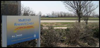 The sign at the MnROAD Research Center is pictured with the test road and traffic in the background.