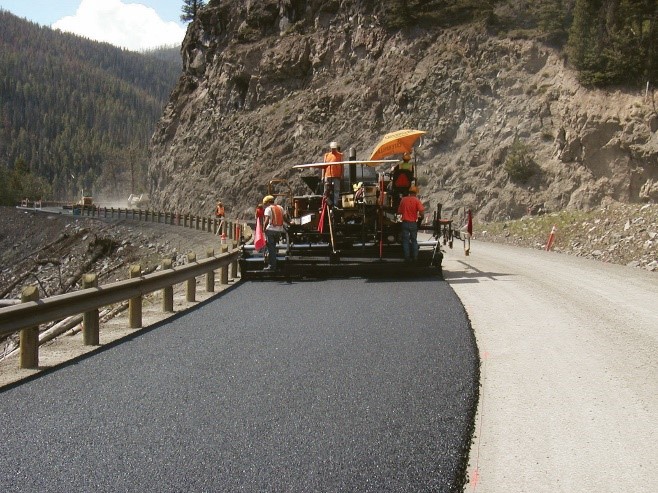 Asphalt is being paved on a roadway alongside a guardrail. The other side of the road is at the base of a steep, rocky cliff.