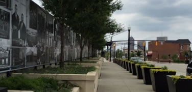 A photo of the cultural wall and side walk which has murals depicting famous people from the area, trees, and planter boxes.