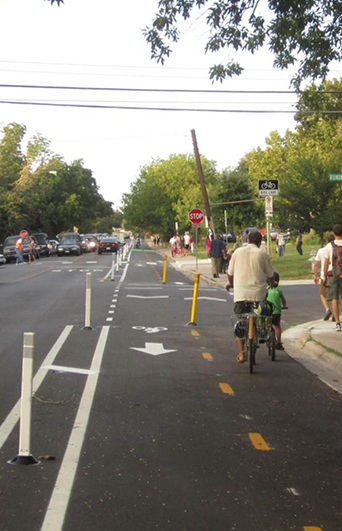 Cyclists and pedestrians are using a bike lane that is sectioned off from the main road by street markings and safety posts.