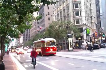 Busy city street corner depicting tall buildings and cars, buses, and bicyclists traveling along the street.