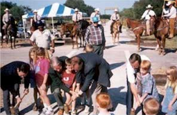 A group of men and children using sledgehammers along a section of walking trail. Behind them are a line of men on horseback.