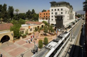 Image shows higher density, mixed use development surrounding a plaza located next to a commuter rail train and station.  Many pedestrians are in the plaza after just apparently disembarking from the train