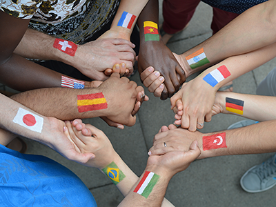 A high-angle view of a diverse group holding hands in a circle, with various national flags painted on their forearms.