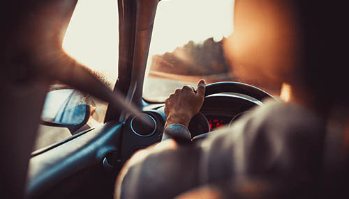 A view from the back seat of a car looking over the driver's shoulder. The driver's hands are gripping the steering wheel, and warm sunlight from a sunset is streaming through the windshield, creating a bright lens flare.