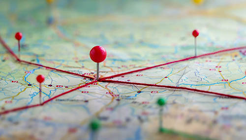 A close-up of a map with several red pushpins connected by red strings, featuring one central pin in sharp focus against a blurred background
