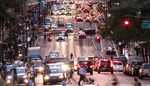 A high-angle, wide shot of a busy city avenue, likely in New York City, packed with traffic and pedestrians in the late afternoon. Warm sunlight reflects off the asphalt, illuminating numerous yellow taxis and cars with headlights on, while crowds of people cross the intersections and fill the sidewalks during the evening commute.