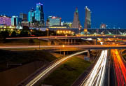 Long exposure photo of a city skyline at night with interstate roads leading to and from the city. Car headlights and brake lights form continuous trails along the roads.