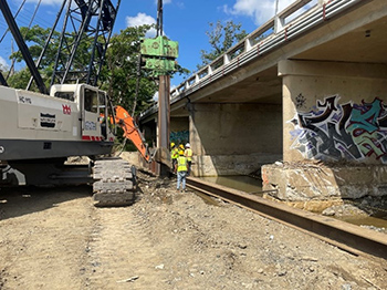 Workers next to heavy machinery on one side of the bottom of a bridge.