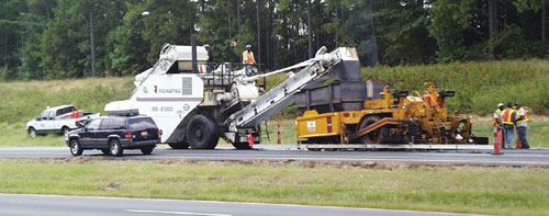 Truck lays down asphalt on road. A worker is standing on the truck. Three workers are in the background on the side of the road.