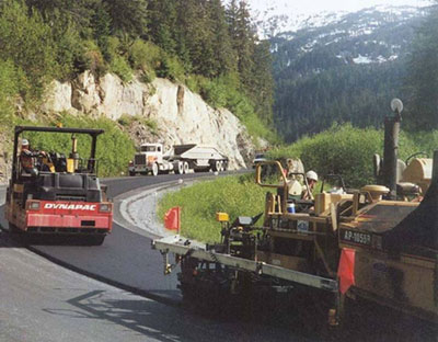 A newly paved asphalt pavement. Two pavers are on the road. A truck is in the background.