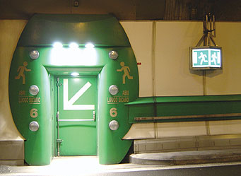 Figure 5. Photo. Mont Blanc Tunnel. A door with a white arrow indicates a tunnel emergency escape route in the Mont Blanc tunnel on the French-Italian border. A sign next to the door shows two white-colored running figures.