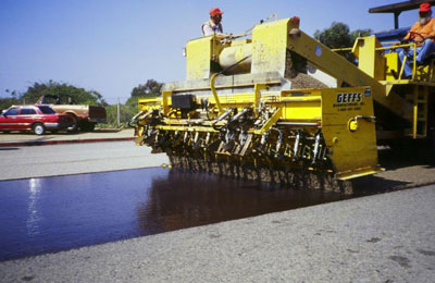 The photo shows a chip seal being applied to a pavement.