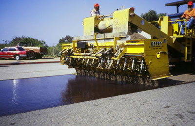 The photo shows a chip seal being applied to a pavement.