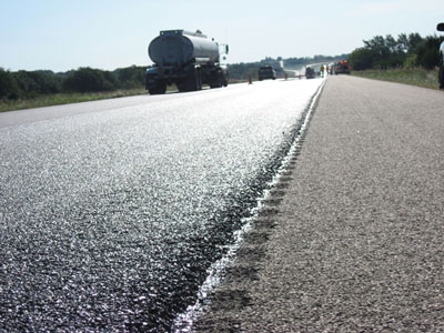 Photo. An ultrathin bonded wearing course on I-10 near San Antonio, TX. The photo shows a close-up view of the asphalt pavement. A tanker truck and two smaller trucks are in the background.