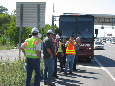 Photo. Team members stop on I-10 in San Antonio, TX. Ten scanning tour team members stand on the side of I-10, in front of the team bus. Traffic is traveling by on I-10.