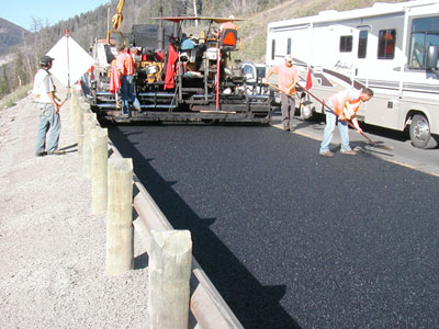 Figure 5. Photo. A close-up of a paver and four workers as warm-mix asphalt is placed on the East Entrance Road in Yellowstone National Park.