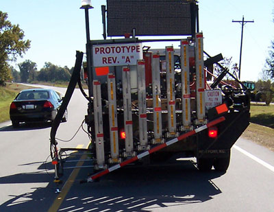 Figure1. Photo. The Automated Roadway Pavement Marker Placement System travels down a roadway, installing raised reflective pavement markers. The device is mounted on a truck. A car can be seen in the lane next to the truck.