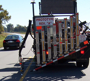 Figure 3. Photo. The Automated Roadway Pavement Marker System travels down a roadway, installing raised reflective pavement markers. The device is mounted on a truck. A car can be seen in the lane next to the truck.