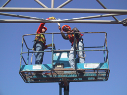 Figure 3. Photo. Two workers on a raised work platform apply fiber-reinforced polymer composite wraps to an overhead sign structure.
