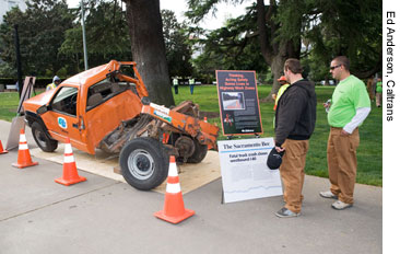 Figure 4. Photo. The wreckage of a Caltrans pickup truck that was struck by a truck on I-80 this year is displayed at the National Work Zone Awareness Week kickoff event in Sacramento, CA. Two men are looking at the display.