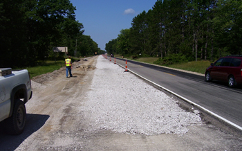 Figure 2. Photo. A lane of roadway on M-115 in Clare County, Michigan, during the paving process. A roadway worker is visible on the side of the road.
