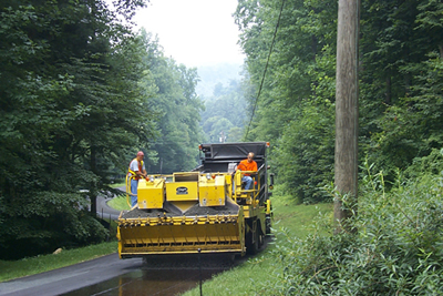 Figure 6. Photo. Two workers on a chip spreader place a chip seal on a roadway.