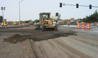 Photo. Road construction equipment prepares a road for paving. Soil is turned up in the forefront of the photo. To the right, a roadway worker in an orange safety vest and hard hat is visible.