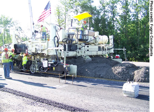 A view of paving equipment being used for long-life concrete pavement construction along a section of I-90 near Syracuse, NY. Three workers are visible.