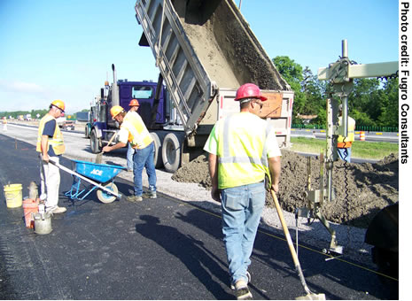 Long-life concrete pavement construction along a section of I-90 near Syracuse, NY. Five workers are visible. A dump truck is unloading materials.