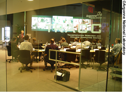 A view of the Transportation Research and Analysis Computer Center at the Argonne National Laboratory in Chicago, IL. Students are sitting at tables in the Center, watching a presentation on projection screens.
