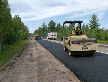 Photo. The Michigan Department of Transportation performs asphalt paving on a two-lane road on M-115 in Clare County, Michigan. A paver with an operator is visible in the foreground, with traffic in the background traveling in the other lane.
