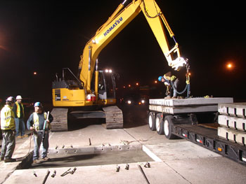 Figure 5. Photo. Precast concrete pavement slabs are used to repair a section of I-295 in New Jersey in 2008. A bulldozer is picking up a concrete slab to lift it into place on the roadway. A construction worker stands in the forefront of the photo.