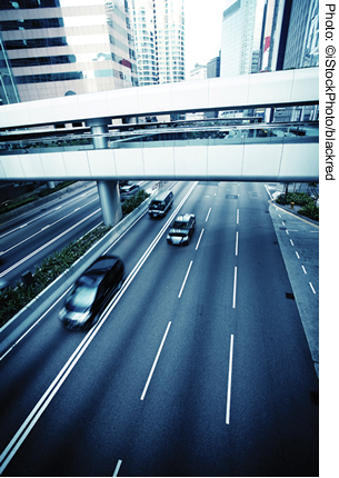 Traffic moving on a four-lane urban roadway. The roadway travels under an overpass. High rise buildings are visible in the background.