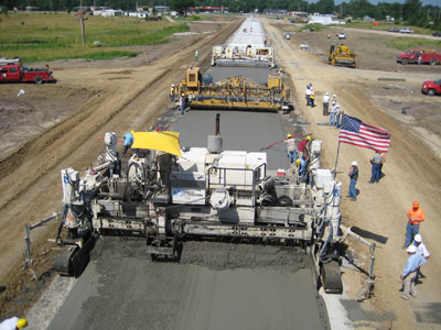 Concrete paving being performed on a roadway. Paving equipment and workers are visible.