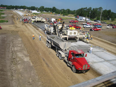 A two-lift concrete pavement test section under construction in Pleasanton, KS, in June 2008, on U.S. 69 and East 1100 Road. Paving equipment, dump trucks, and workers are visible.