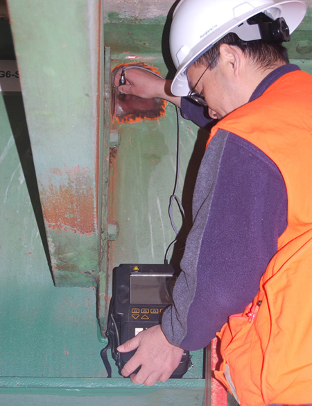 A close-up of FHWA Nondestructive Evaluation Center researcher Rongtang Liu, who is using the Eddy Current system to inspect a weld on a stiffener. Liu wears an orange vest and white hard hat.