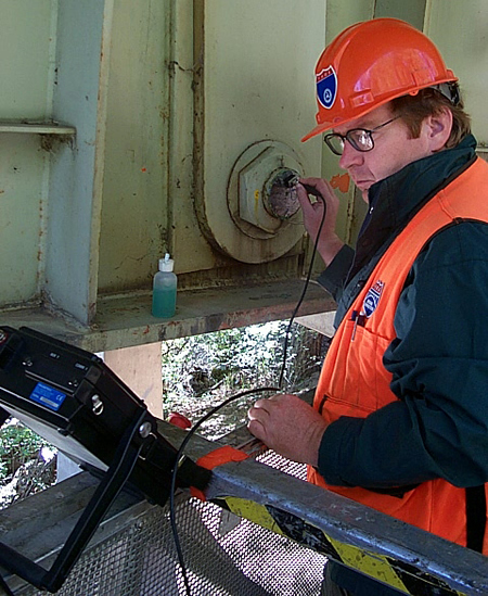 A close-up of bridge inspector Marcus Miller of FHWA's Eastern Federal Lands Highway Division office, who is using the Ultrasonic system to inspect a bridge pin. Miller wears an orange hard hat and vest as he views the system.