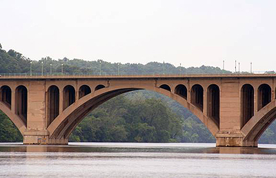 A ground-level view of a bridge arch spanning a river. A wooded area is in the background.