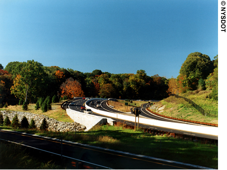 Vehicles traveling on a four-lane, divided roadway constructed with asphalt pavement. Trees and fall foliage frame the roadway in the background.