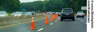 A view from the center lane as traffic flows along a three-lane highway. The left lane is closed off by traffic cones. Jersey barriers separate the three lanes of traffic flowing in the opposite direction.