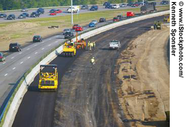 A view looking down over a highway work zone as repaving work is done. Traffic continues to flow in the left-hand lanes of the highway. Paving equipment and five workers are visible.