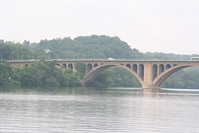 A view from a distance of traffic traveling across a bridge, with water in the foreground. Wooded areas are in the background.